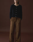 A woman with straight brown hair tucked behind her ears stands against a dark brown backdrop, hands in her pockets, wearing the Mia Fratino Cashmere Sienna Cardi and high-waisted, wide-leg brown pants.