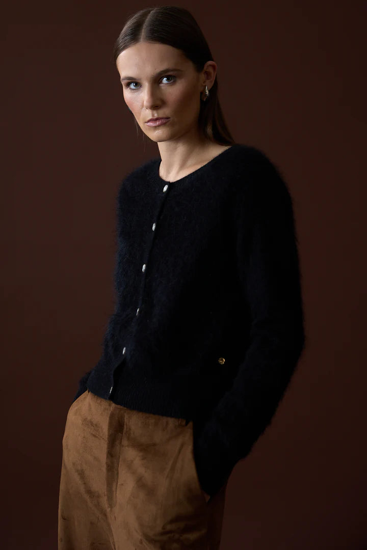 A woman with straight brown hair models the Mia Fratino Cashmere Sienna Cardi, a slim fit fuzzy black button-front cardigan, paired with brown pants. She stands with hands in pockets against a plain brown background.