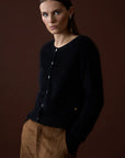 A woman with straight brown hair models the Mia Fratino Cashmere Sienna Cardi, a slim fit fuzzy black button-front cardigan, paired with brown pants. She stands with hands in pockets against a plain brown background.