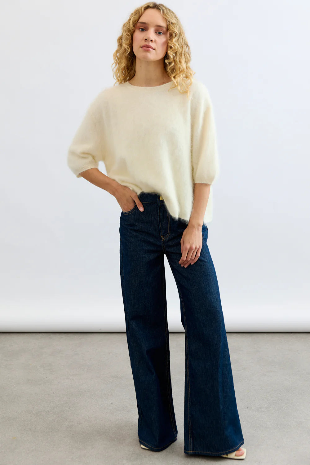 A person with long curly blonde hair wears the Mia Fratino Cashmere Erika Tee and high-waisted wide-leg blue jeans, posing against a plain light background with one hand on their hip.