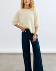 A person with long curly blonde hair wears the Mia Fratino Cashmere Erika Tee and high-waisted wide-leg blue jeans, posing against a plain light background with one hand on their hip.
