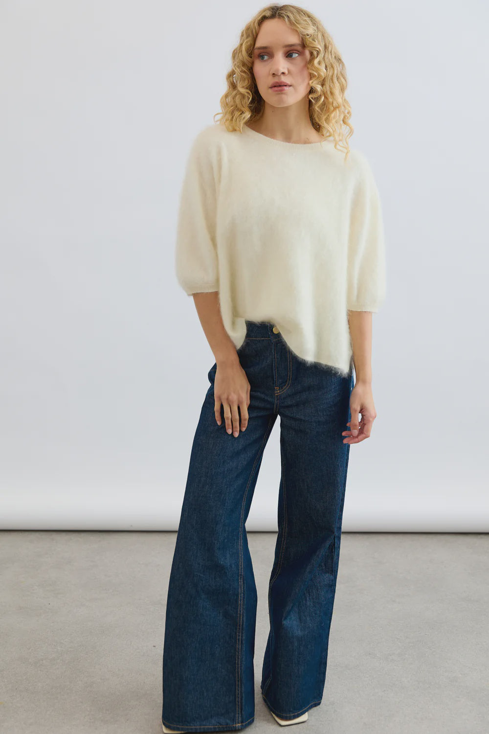 A person with curly blonde hair wears the Erika Tee by Mia Fratino Cashmere and high-waisted wide-leg blue jeans, standing on a light gray floor against a plain white background.