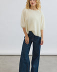 A person with curly blonde hair wears the Erika Tee by Mia Fratino Cashmere and high-waisted wide-leg blue jeans, standing on a light gray floor against a plain white background.