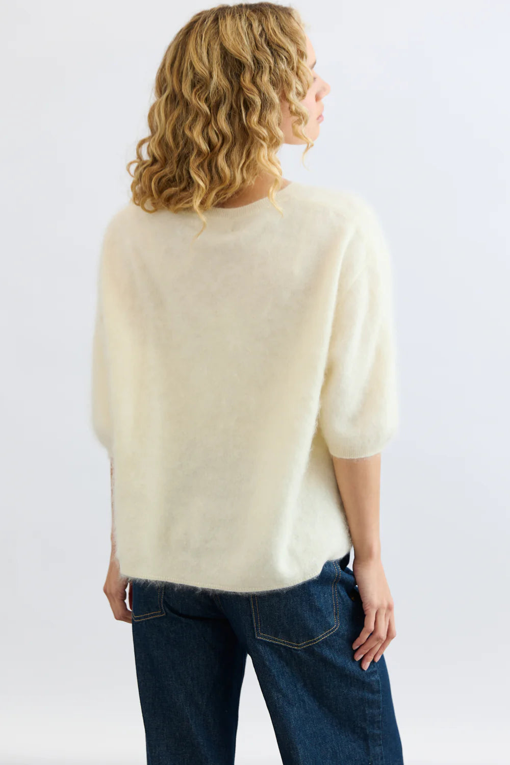 A person with curly blonde hair is seen from the back, wearing the Mia Fratino Cashmere Erika Tee—a fuzzy, brushed cashmere short-sleeve sweater—with blue jeans against a plain light background.