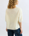 A person with curly blonde hair is seen from the back, wearing the Mia Fratino Cashmere Erika Tee—a fuzzy, brushed cashmere short-sleeve sweater—with blue jeans against a plain light background.