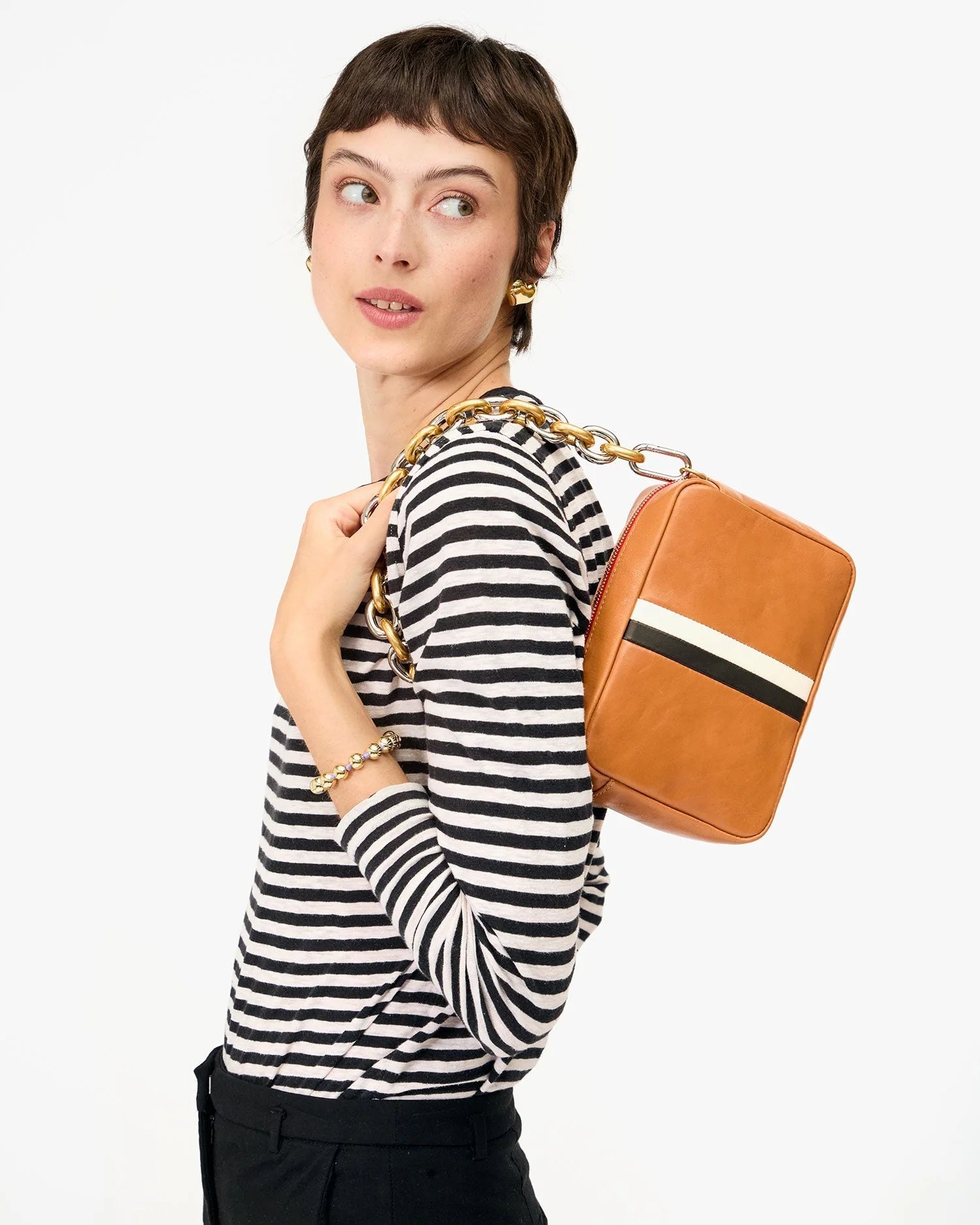 A woman in a black-and-white striped long-sleeve shirt and black pants holds the Clare Vivier Midi Sac S25, a handwoven leather rectangular crossbody with a gold chain strap, resting it on her shoulder and looking to the side.