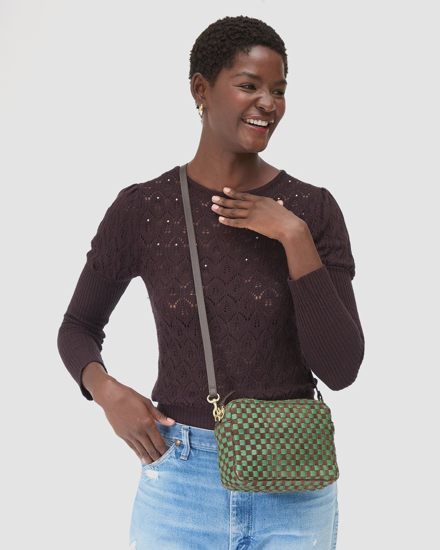 A smiling woman in a dark, textured long-sleeve top and light blue jeans holds the Clare Vivier Midi Sac 26, a handwoven green and brown checkered leather crossbody bag, standing against a plain white background.