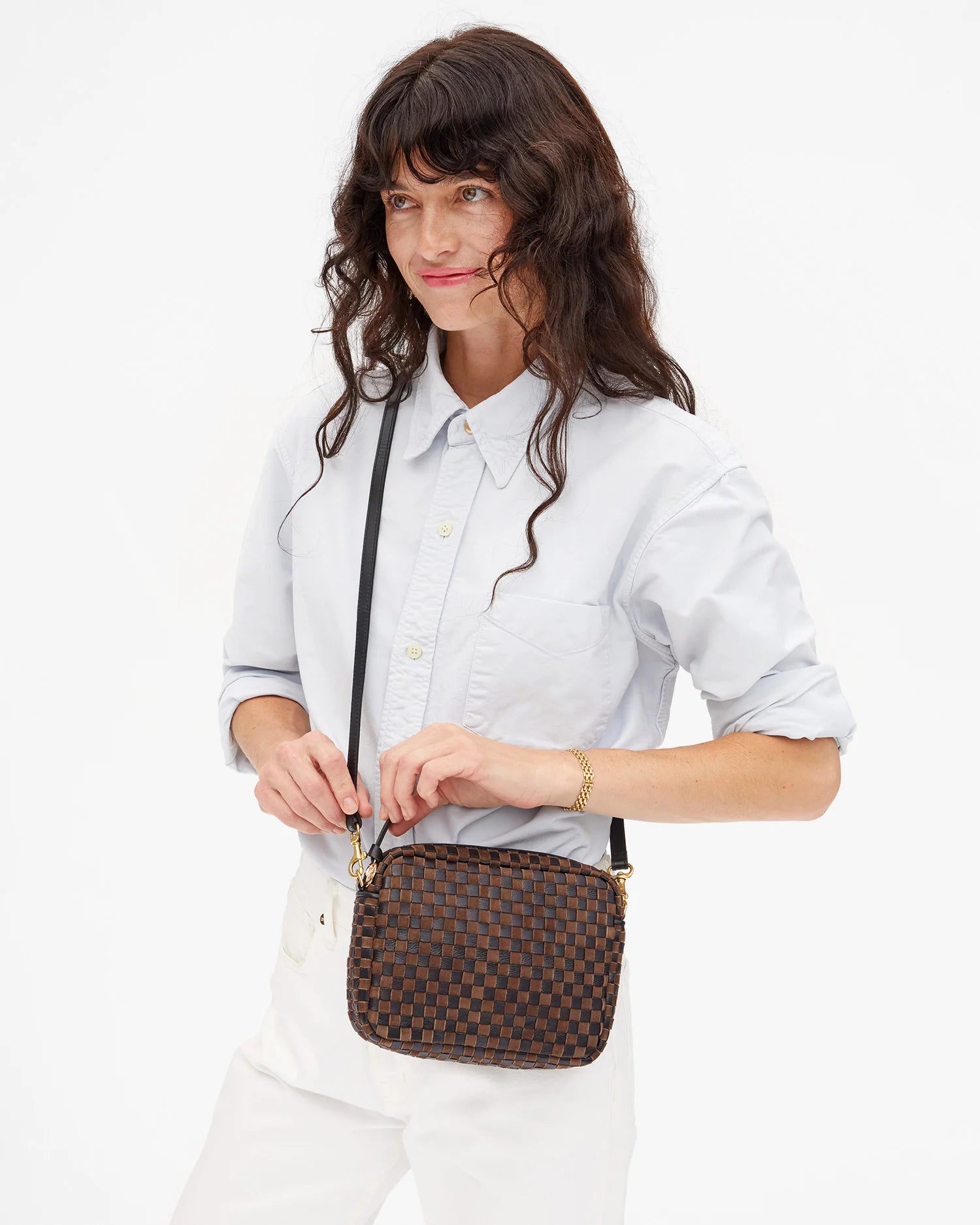 A woman with long wavy brown hair wears a light blue button-up shirt and white pants, holding the Clare Vivier Midi Sac S25 crossbody bag with a black strap against a plain white background.