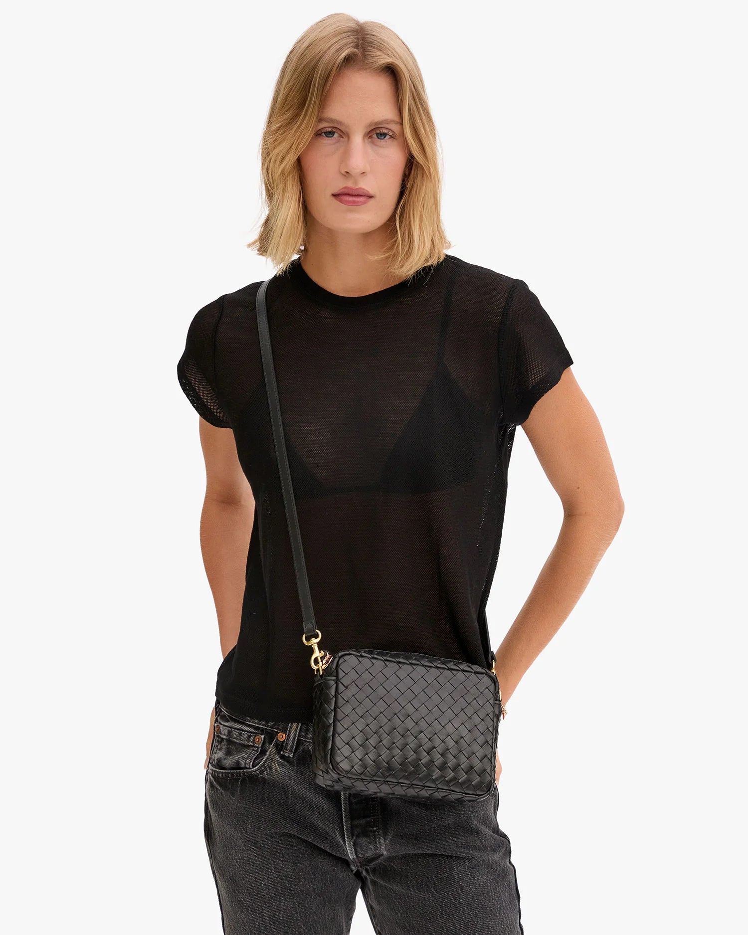 A person with shoulder-length blonde hair wears a sheer black t-shirt, dark jeans, and carries the Clare Vivier Midi Sac S25 crossbody bag with a woven pattern, standing against a plain white background.