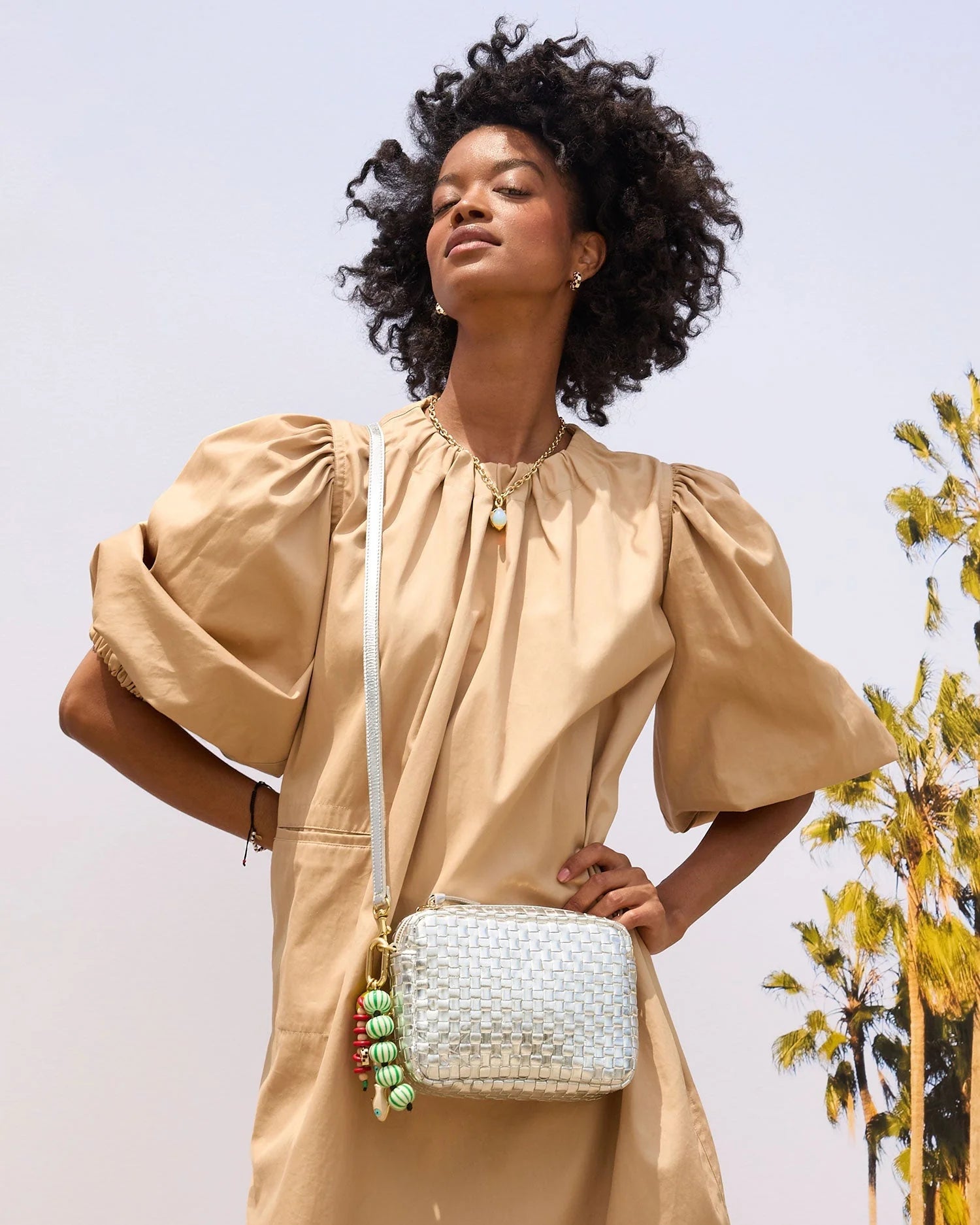 A woman in a beige puff-sleeve dress poses outdoors with the Clare Vivier Midi Sac S25, a chic handwoven leather crossbody bag. Palm trees and a clear sky are behind her. She has curly hair and wears layered necklaces.