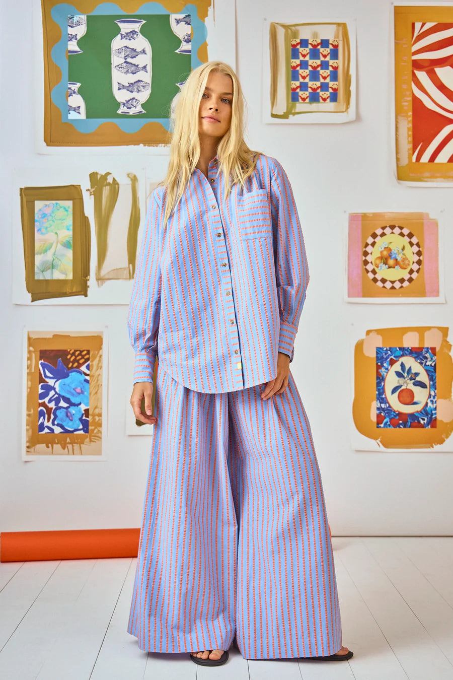 A person with long blonde hair stands in front of a vibrant abstract art wall, wearing a Hunter Bell NYC Beach Stripe button-up paired with Dasha Pant. The scene exudes a bright and artistic vibe.