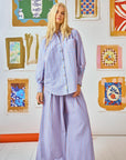 A person with long blonde hair stands in front of a vibrant abstract art wall, wearing a Hunter Bell NYC Beach Stripe button-up paired with Dasha Pant. The scene exudes a bright and artistic vibe.