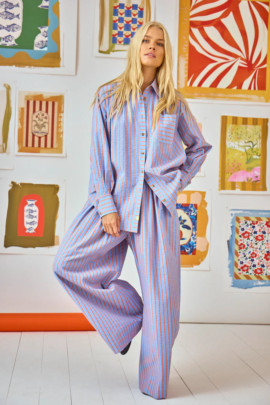 A person with long blonde hair wears the Hunter Bell NYC Dasha Pant and a matching beach stripe shirt, standing in a room filled with colorful, abstract framed artworks. The floor is white, and there's a touch of something orange nearby.