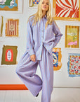 A person with long blonde hair wears the Hunter Bell NYC Dasha Pant and a matching beach stripe shirt, standing in a room filled with colorful, abstract framed artworks. The floor is white, and there's a touch of something orange nearby.