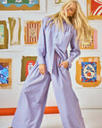 A person with long blonde hair stands in a room adorned with framed art, wearing a blue and red Hunter Bell NYC Miles Shirt. They gaze downward, one hand tucked into the pocket of their relaxed-fit, wide pants.