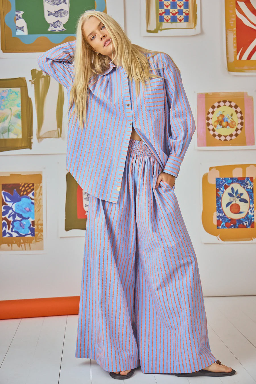 A person with long blond hair wears a matching oversized shirt and Dasha Pant from Hunter Bell NYC, featuring a stylish smocked waistband. The Beach Stripe shirt has orange and blue stripes. They stand before a wall adorned with colorful framed artwork on a pristine white floor.