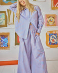 A person with long blond hair wears a matching oversized shirt and Dasha Pant from Hunter Bell NYC, featuring a stylish smocked waistband. The Beach Stripe shirt has orange and blue stripes. They stand before a wall adorned with colorful framed artwork on a pristine white floor.