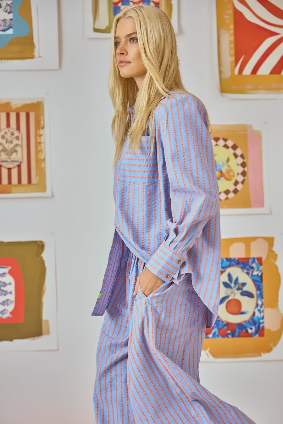 A person with long blonde hair wearing Hunter Bell NYC's Miles Shirt in blue and orange Beach Stripe (100% cotton) stands in profile, hands in pockets, against a wall adorned with colorful abstract art.