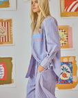 A person with long blonde hair wearing Hunter Bell NYC's Miles Shirt in blue and orange Beach Stripe (100% cotton) stands in profile, hands in pockets, against a wall adorned with colorful abstract art.