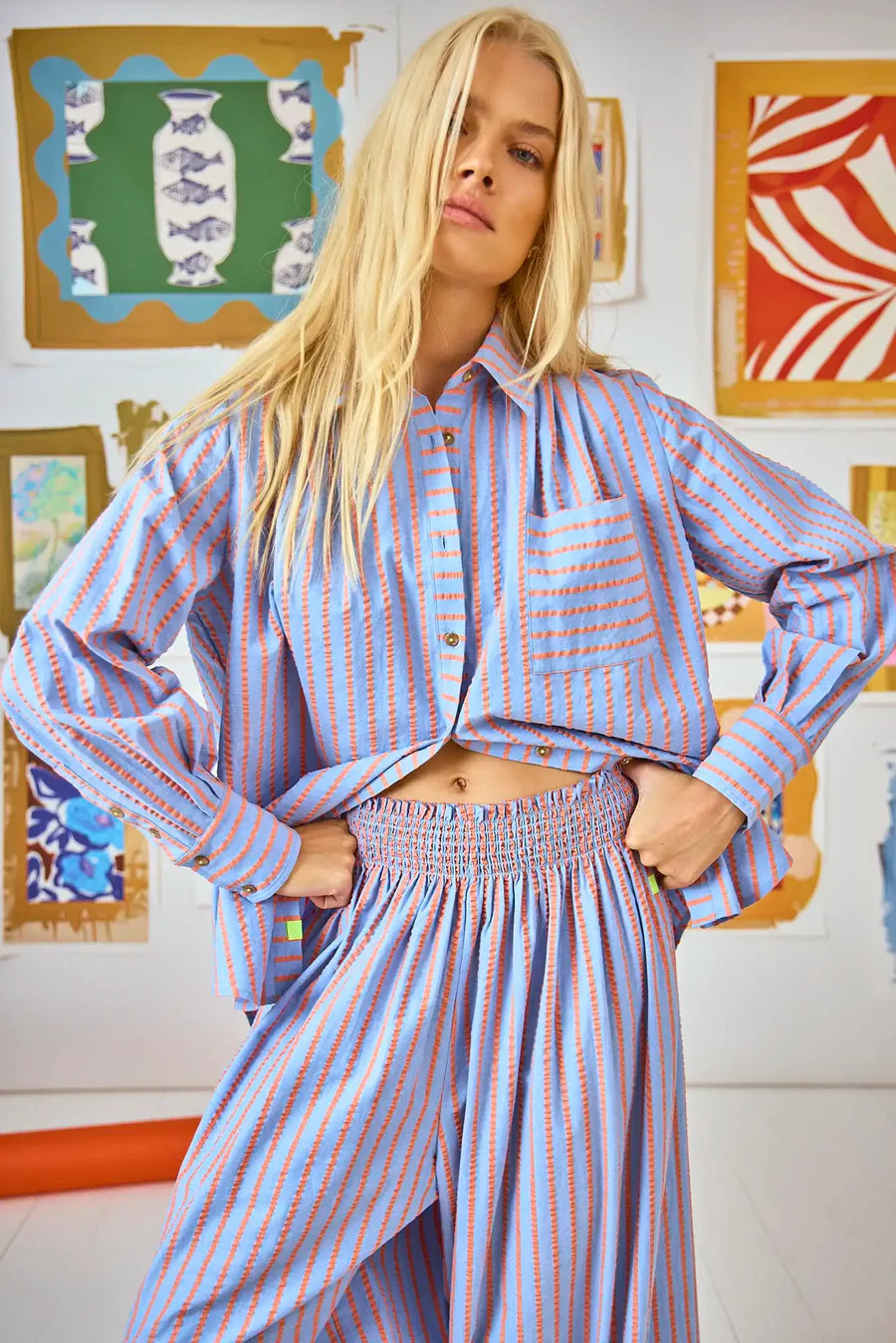 A person with long blonde hair is wearing the relaxed fit Miles Shirt by Hunter Bell NYC, featuring blue and orange stripes and a button front. They are indoors, standing in front of a wall decorated with colorful framed artwork.