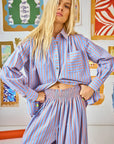 A person with long blonde hair is wearing the relaxed fit Miles Shirt by Hunter Bell NYC, featuring blue and orange stripes and a button front. They are indoors, standing in front of a wall decorated with colorful framed artwork.