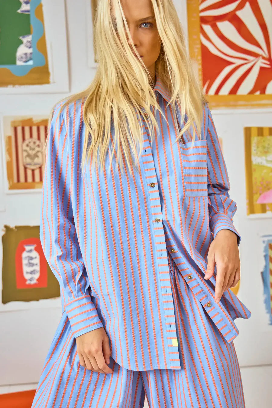 A person with long blonde hair is wearing the Miles Shirt by Hunter Bell NYC, featuring a relaxed fit with blue and red beach stripes, paired with matching pants. They stand before a wall adorned with colorful framed artworks, exuding laid-back elegance.