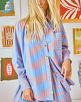 A person with long blonde hair is wearing the Miles Shirt by Hunter Bell NYC, featuring a relaxed fit with blue and red beach stripes, paired with matching pants. They stand before a wall adorned with colorful framed artworks, exuding laid-back elegance.