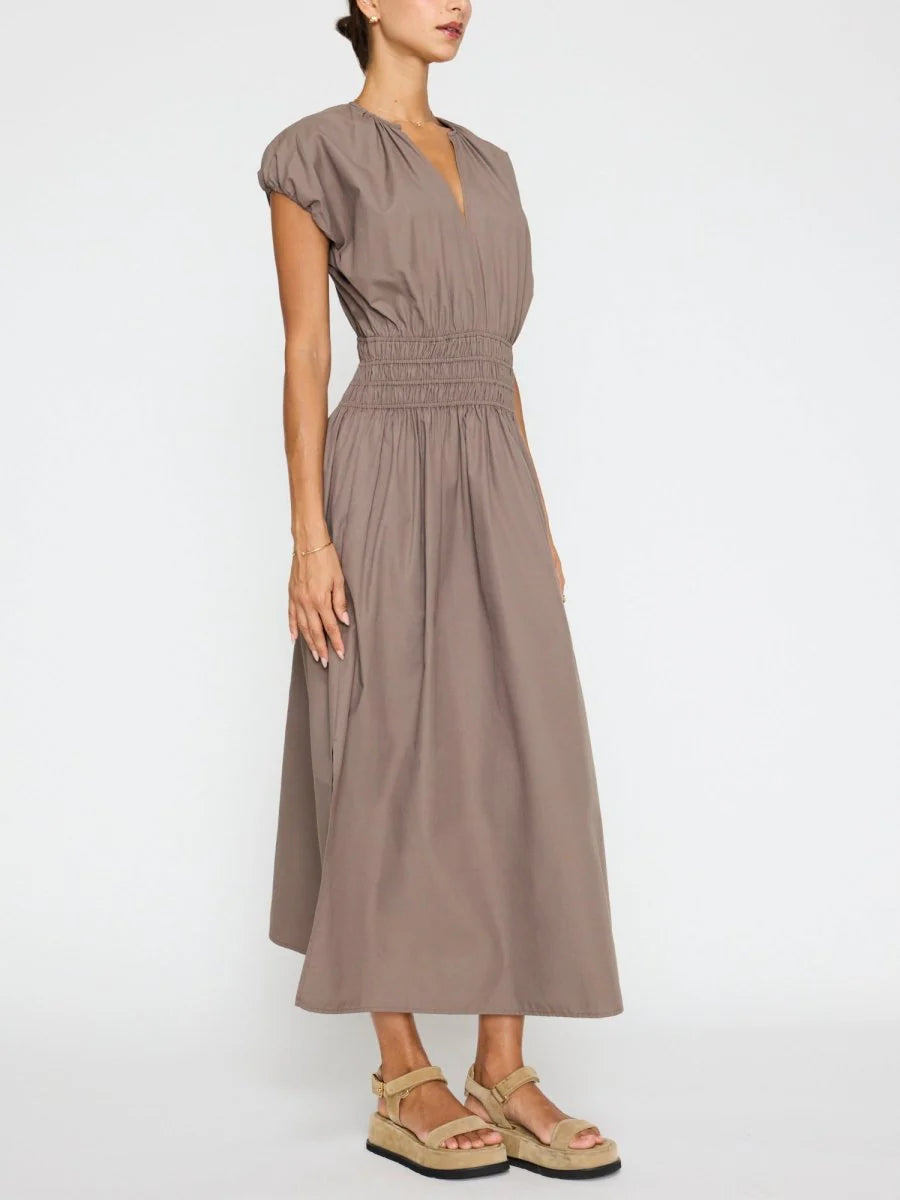 A woman wears the BROCHU WALKER MINA DRESS, a taupe short-sleeve midi with v-neckline and smocked waist, paired with tan platform sandals, standing against a plain white background.