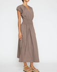 A woman wears the BROCHU WALKER MINA DRESS, a taupe short-sleeve midi with v-neckline and smocked waist, paired with tan platform sandals, standing against a plain white background.