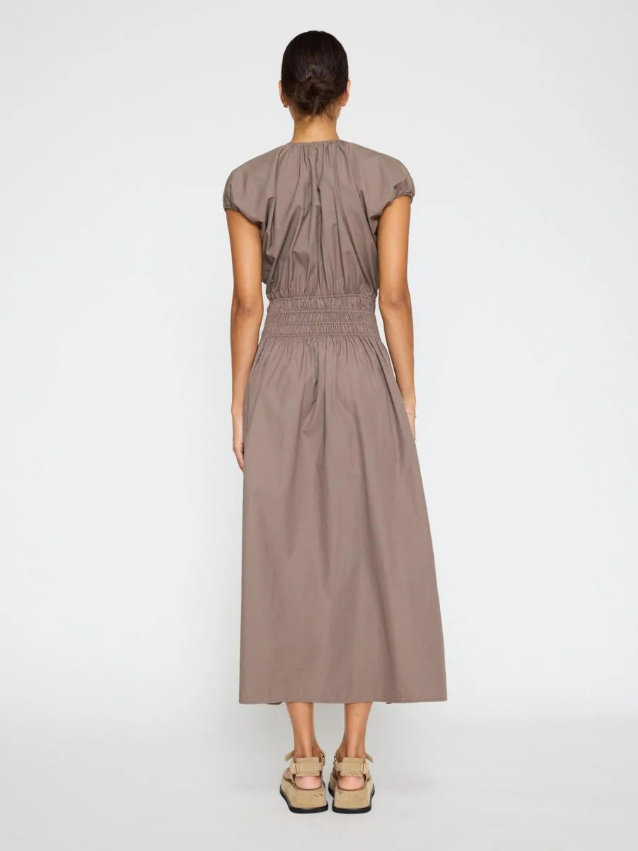 A woman stands with her back turned, wearing the BROCHU WALKER MINA DRESS in taupe—an ankle-length midi with a gathered waist—her hair tied back and styled with beige platform sandals against a plain white background.
