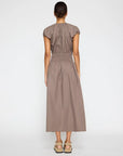A woman stands with her back turned, wearing the BROCHU WALKER MINA DRESS in taupe—an ankle-length midi with a gathered waist—her hair tied back and styled with beige platform sandals against a plain white background.