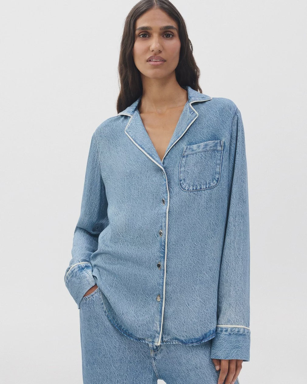 A woman with long dark hair wears the Rag & Bone miramar slink long sleeve lounge shirt and matching denim pants, standing against a plain white background, one hand in her pocket.