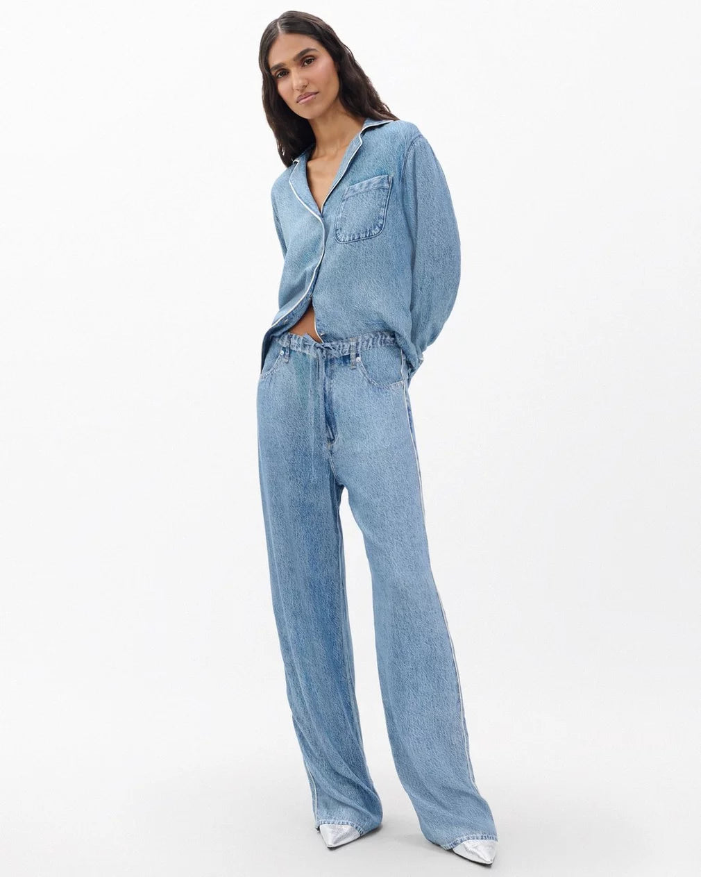 A woman with long dark hair wears Rag & Bone’s miramar slink lounge pant in a trompe l’oeil denim print with a matching shirt, both crafted from soft loungewear fabric, against a plain white background. She finishes the look with pointed silver shoes.