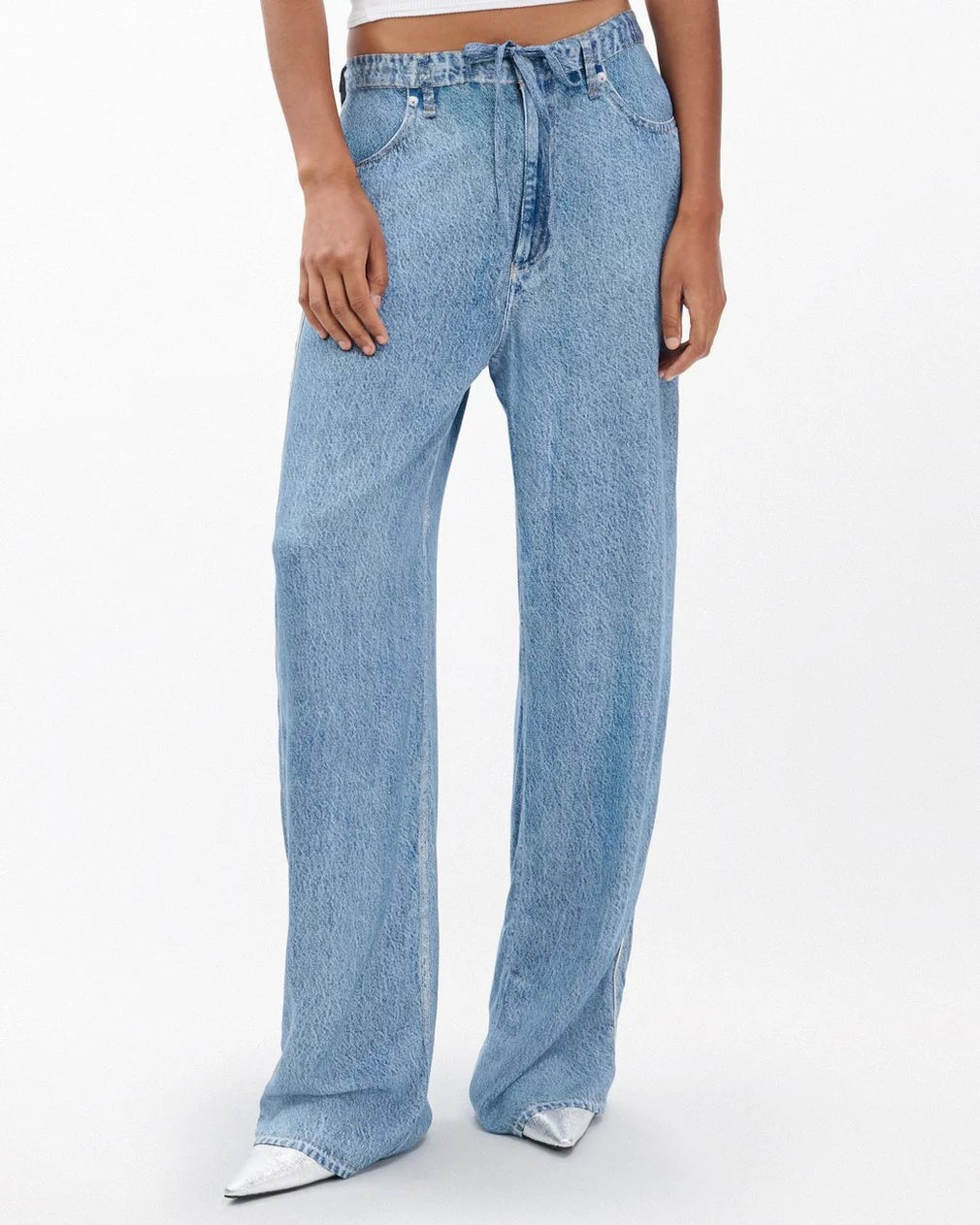 A person wears Rag & Bone's miramar slink lounge pant—high-waisted, loose-fit trompe l'oeil denim with a tie belt—paired with white pointed shoes. Only the lower torso and legs show against a plain background.