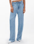 A person wears Rag & Bone's miramar slink lounge pant—high-waisted, loose-fit trompe l'oeil denim with a tie belt—paired with white pointed shoes. Only the lower torso and legs show against a plain background.