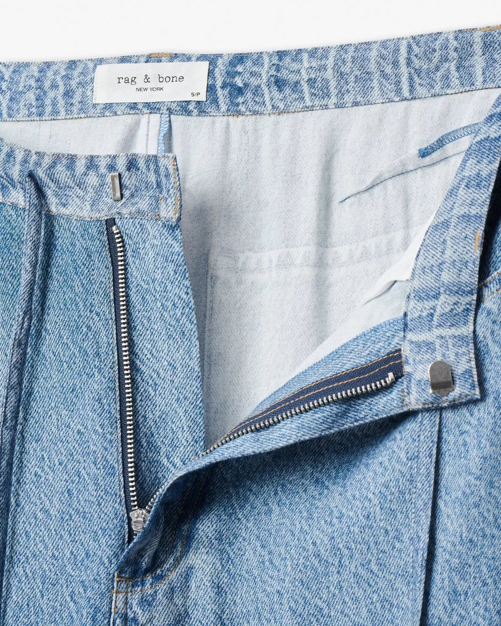 Close-up of light blue Rag &amp; Bone miramar slink lounge pant with undone waistband, zipper, and metal button, revealing white interior lining and visible brand label inside waistband.