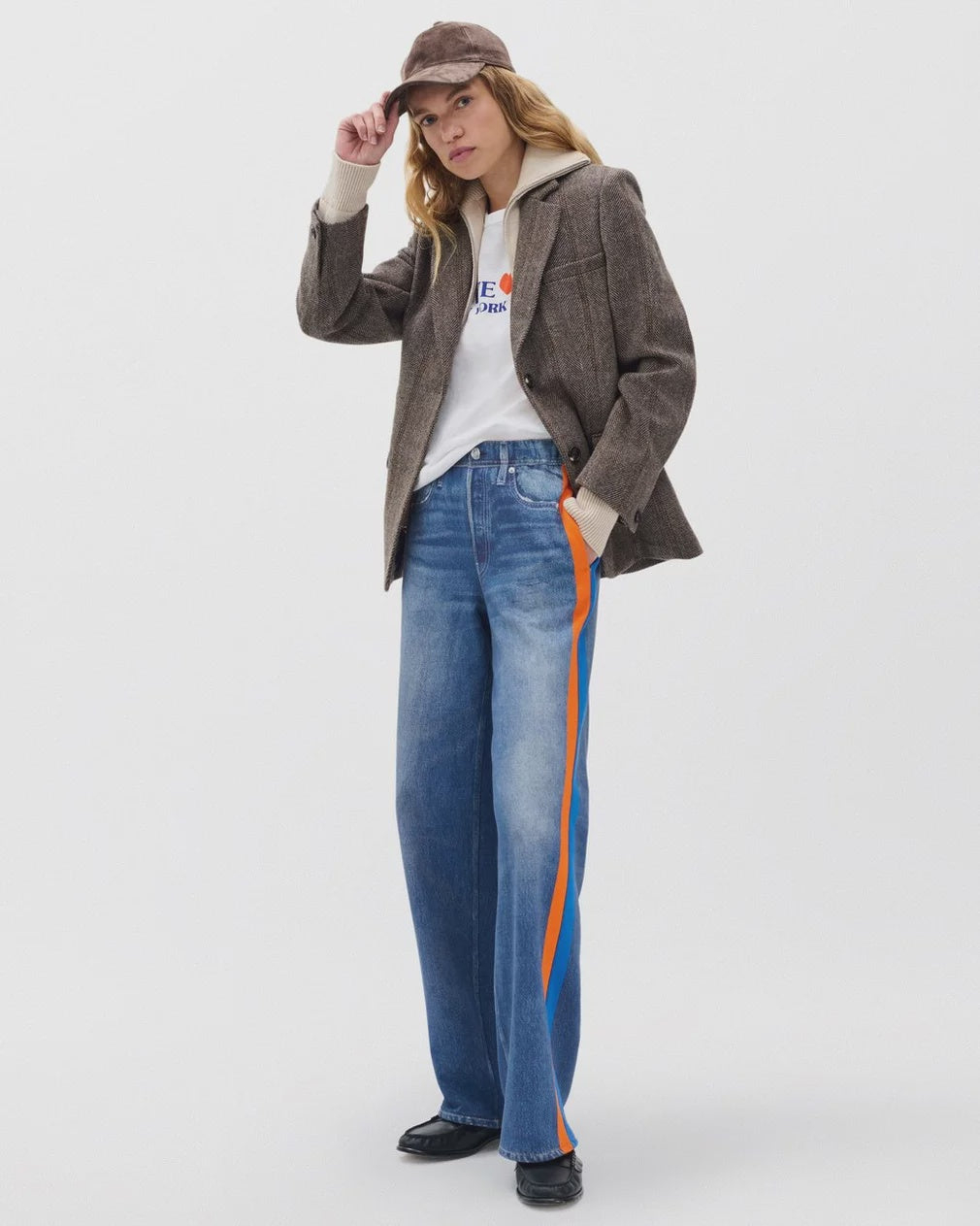 A person stands against a plain white background wearing Rag & Bone’s miramar terry track pant pull on, a brown cap, brown blazer, white hoodie, graphic T-shirt, and black shoes.