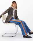 A person wears a brown blazer, graphic white tee, Rag & Bone miramar terry track pant pull on, black loafers, and a brown cap while seated on a modern white chair against a plain background.