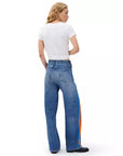 A woman with long blonde hair wears a white cropped t-shirt and Rag & Bone miramar terry track pant pull ons, limited edition denim style with orange and blue side stripes, standing and facing away from the camera.