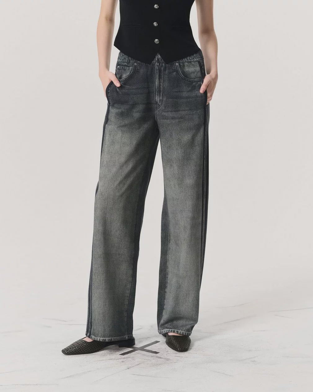 A person stands against a plain light background with hands in pockets, wearing Rag & Bone’s Miramar Wide Leg Track Pant in faded black denim look, a black sleeveless buttoned top, and black pointed-toe flats.