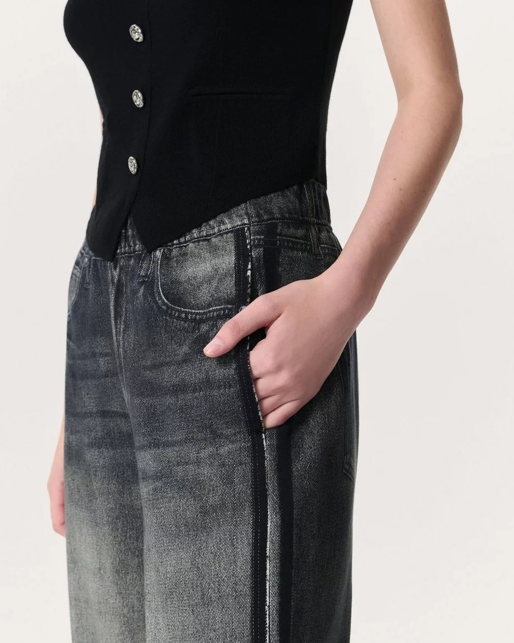 A person wears Rag & Bone's Miramar Wide Leg Track Pant and a sleeveless black buttoned top, standing with one hand in their pocket against a light background.