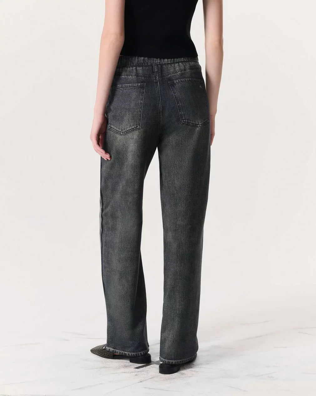 A person stands facing away from the camera, wearing a black sleeveless top and Rag & Bone Miramar Wide Leg Track Pants in faded black denim with back pockets against a plain light background.