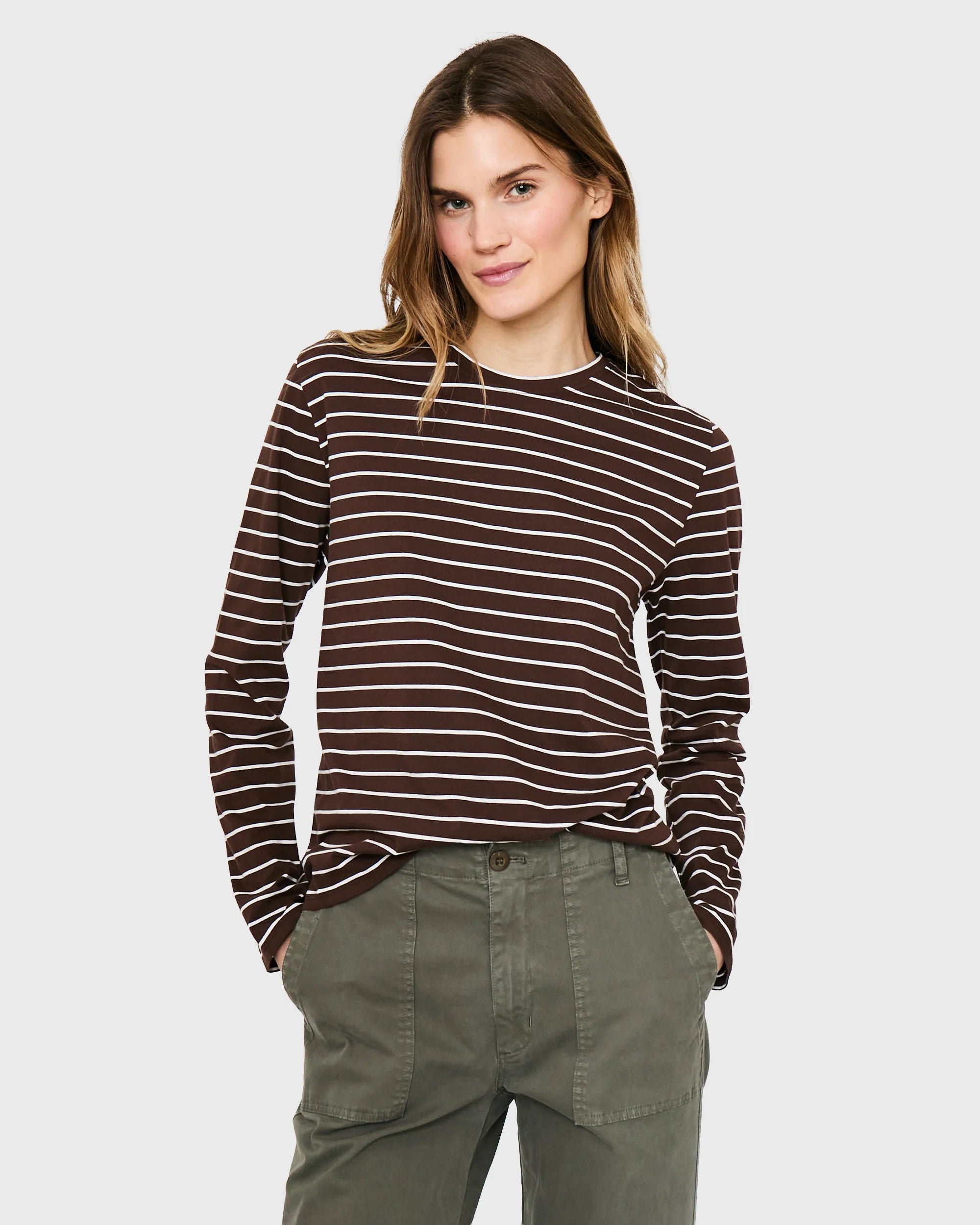A person with light brown hair models Kule's The Modern Long, a brown and white striped long sleeve tee, paired with olive green pants, standing hands-in-pockets against a plain light gray background.