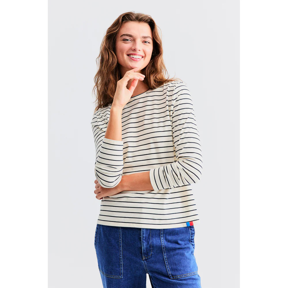 A smiling person with wavy hair models "The Modern Long" by Kule, a black and white striped long-sleeve shirt crafted from Portuguese cotton, paired with blue jeans, against a light gray background. One hand rests on their chin.