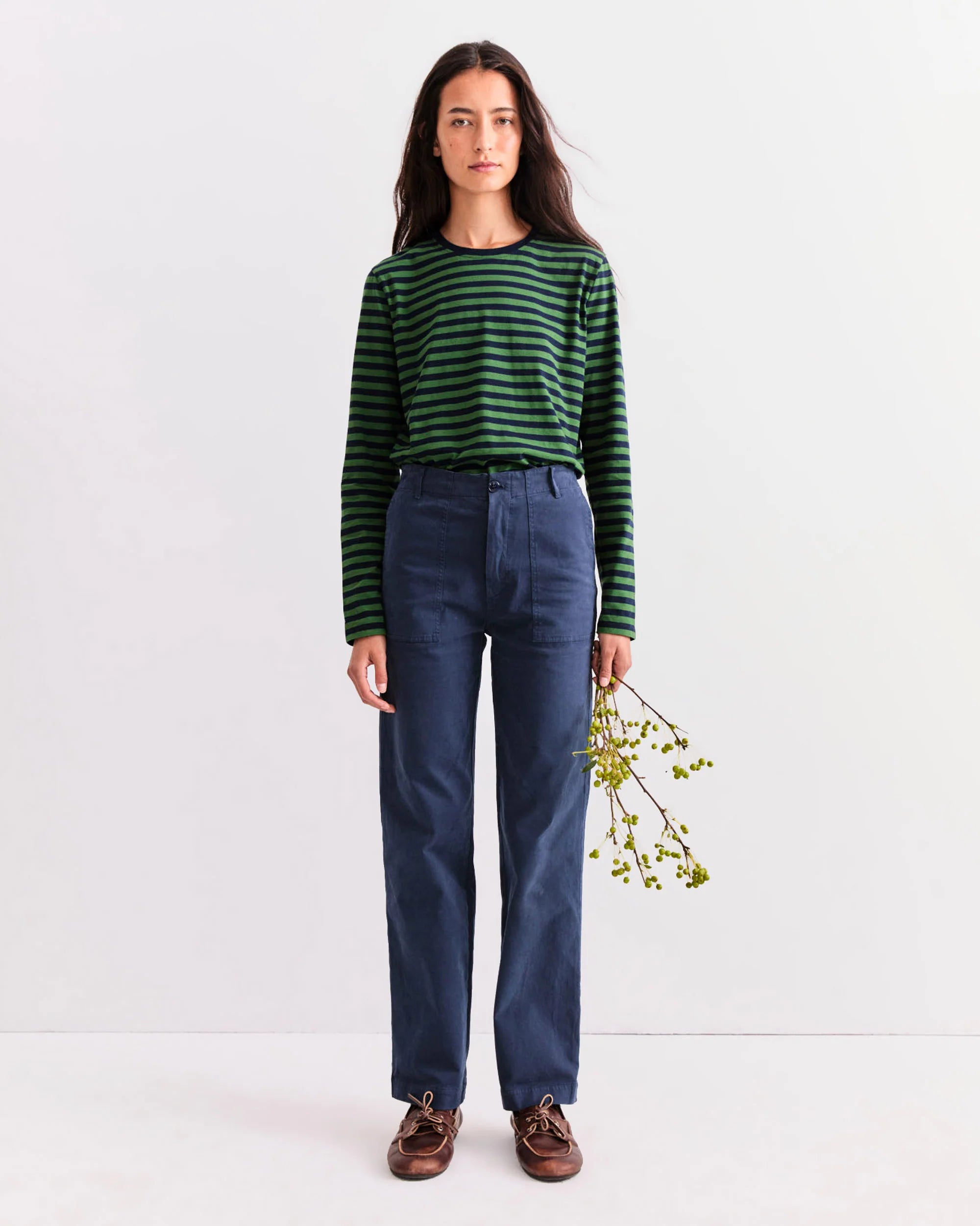 A person with long brown hair stands against a white wall, wearing Kule’s The Modern Long— a green and black striped long sleeve shirt—paired with high-waisted navy pants and brown shoes, holding a sprig of green foliage in their right hand.