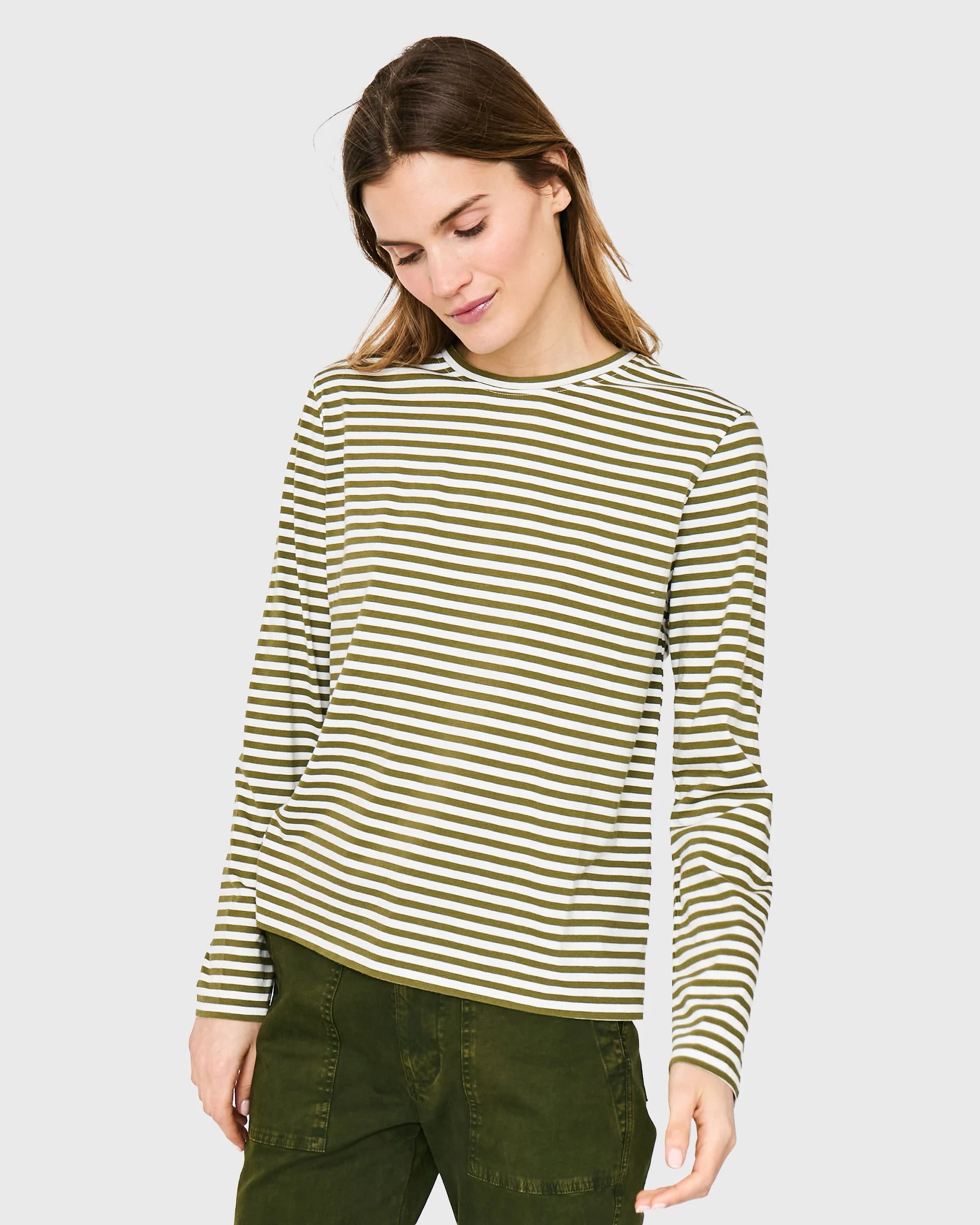 A woman with long brown hair wears Kule's The Modern Long, a green and white striped long sleeve tee, paired with green pants. She looks down and slightly smiles against a plain light gray background.
