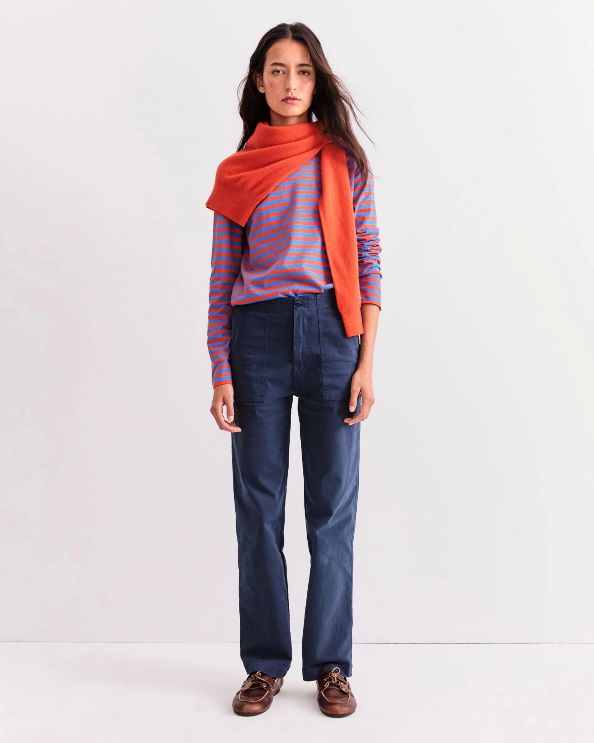 A woman stands against a white background wearing Kule's The Modern Long blue and red striped cotton long sleeve tee, navy pants, brown shoes, and an orange sweater draped over her shoulders.