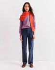 A woman stands against a white background wearing Kule's The Modern Long blue and red striped cotton long sleeve tee, navy pants, brown shoes, and an orange sweater draped over her shoulders.