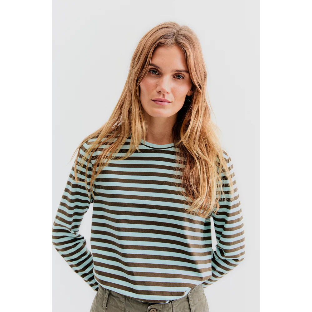 A person with long hair wearing Kule's The Modern Long, a lightweight Portuguese cotton striped long-sleeve shirt, stands against a plain background.