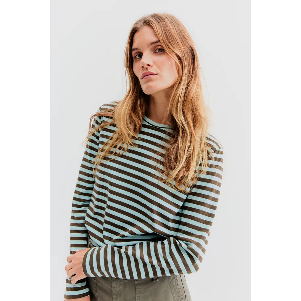 A person with long, wavy hair is wearing The Modern Long by Kule, a Portuguese cotton striped long-sleeve shirt, paired with green pants against a plain white background.